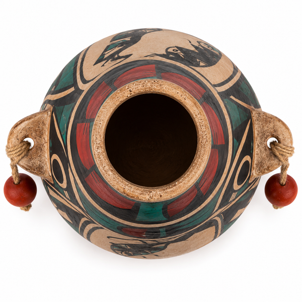 A hand-painted ceramic pot with a circular opening, featuring a tan clay base decorated with red, green, and black geometric and organic patterns. It has two side handles made from natural wood, each attached to a cord loop holding a small red bead.